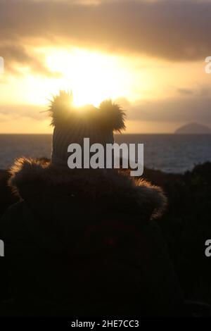 Dunure, Ayrshire, Schottland, Großbritannien. Eine Frau am Wintertag sitzt mit dem Rücken zur Kamera und schaut den Sonnenuntergang an der Ayrshire Coast an, mit einem warmen Bommel-Hut Stockfoto