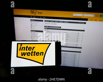 Person, die Mobiltelefon mit Logo des Sportwettenunternehmens Interwetten Ltd. Auf dem Bildschirm vor der Business-Webseite hält. Konzentrieren Sie sich auf die Telefonanzeige. Stockfoto