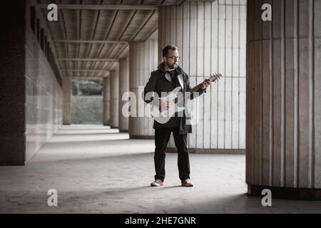 Mann in Schwarz mit Bart, der an einem städtischen Außengelände E-Gitarre spielt. Gitarre, Solo und Street Music Konzept Stockfoto