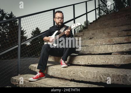 Portrait eines männlichen Gitarristen mit E-Gitarre in schwarzer Kleidung und roten Sneakers auf einer Treppe in der Stadt Stockfoto