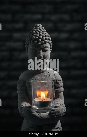 Sitzend in Meditation Buddha Skulptur auf schwarzem Backstein Wand Hintergrund, mit Kerze. Zen- und Meditationskonzept. Stockfoto