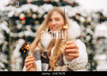 Die Frau in einer Daunenjacke und einem warmen Schal hält Wunderkerzen in der Hand und feiert auf der Straße neben einem Weihnachtsbaum Neujahr oder Weihnachten, ein Wom Stockfoto