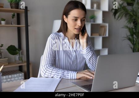 Ernsthafter junger Mitarbeiter, der am Telefon spricht und einen Laptop verwendet Stockfoto