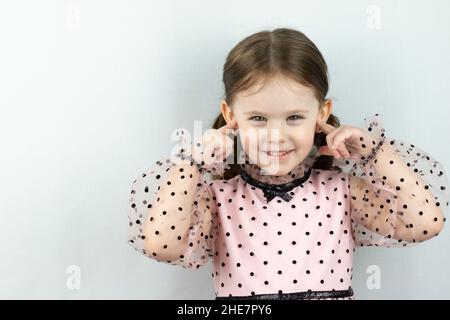 Lächelndes kleines Mädchen mit zwei Pferdeschwänzen in einem Kleid mit Tupfen auf weißem Hintergrund schließt ihre Ohren mit den Fingern. Studio-Foto Stockfoto