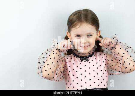 Lächelndes kleines Mädchen mit zwei Pferdeschwänzen in einem Kleid mit Tupfen auf weißem Hintergrund schließt ihre Ohren mit den Fingern. Studio-Foto Stockfoto