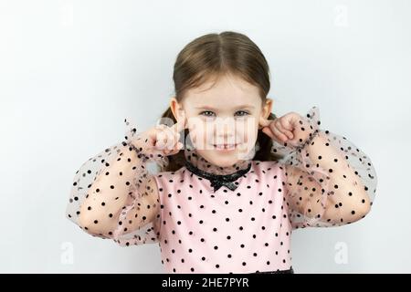 Lächelndes kleines Mädchen mit zwei Pferdeschwänzen in einem Kleid mit Tupfen auf weißem Hintergrund schließt ihre Ohren mit den Fingern. Studio-Foto Stockfoto