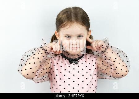 Lächelndes kleines Mädchen mit zwei Pferdeschwänzen in einem Kleid mit Tupfen auf weißem Hintergrund schließt ihre Ohren mit den Fingern. Studio-Foto Stockfoto