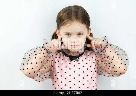 Lächelndes kleines Mädchen mit zwei Pferdeschwänzen in einem Kleid mit Tupfen auf weißem Hintergrund schließt ihre Ohren mit den Fingern. Studio-Foto Stockfoto