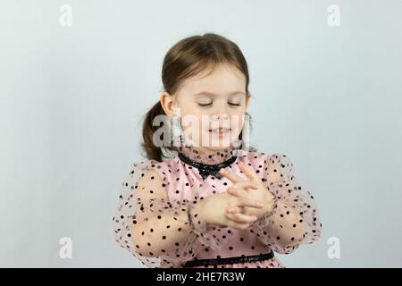 Lächelndes kleines Mädchen mit zwei Pferdeschwänzen in einem Kleid mit Tupfen auf weißem Hintergrund hält beide Hände vor sich. Studio-Foto Stockfoto