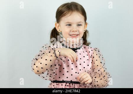 Lächelndes kleines Mädchen mit zwei Pferdeschwänzen in einem Kleid mit Tupfen auf weißem Hintergrund hält beide Hände vor sich. Studio-Foto Stockfoto