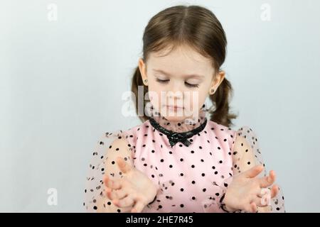 Lächelndes kleines Mädchen mit zwei Pferdeschwänzen in einem Kleid mit Tupfen auf weißem Hintergrund spreizt ihre Hände vor sich. Studio-Foto Stockfoto