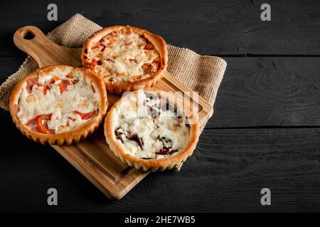 Drei kleine Pizzen auf einem Schneidebrett auf schwarzem Holzhintergrund. Mit einem Kopierbereich. Stockfoto