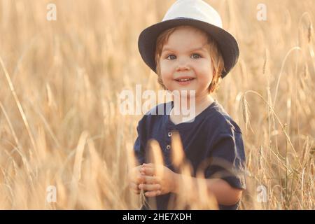 Nahaufnahme eines niedlichen kaukasischen kleinen Jungen im Alter von 2-3 Jahren in einem Strohhut, der an einem Sommertag bei Sonnenuntergang auf einem Weizenfeld läuft und spielt Stockfoto