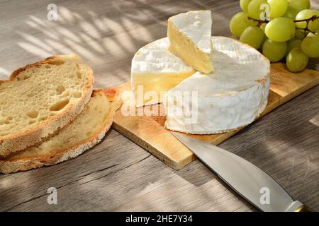 Französischer Camembert der Normandie serviert auf Holzschneidebrett mit Trauben und Brotscheiben. Stockfoto