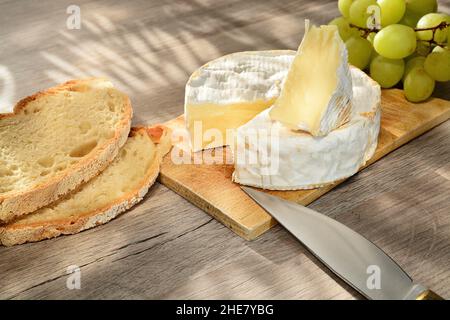 Französischer Camembert der Normandie serviert auf Holzschneidebrett mit Trauben und Brotscheiben. Stockfoto