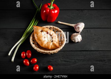 Pie in einem Korbkorb auf schwarzem Holzgrund. Mit einem Kopierraum.Gemüsekomposition - Tomaten, Pfeffer, Zwiebel, Knoblauch. Stockfoto