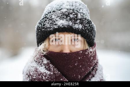 Blonde Frau in Mütze Hut und Nase mit Schal bedeckt schaut auf die Kamera im Winter Schneefall. Nordisches Winterporträt, ruhige und ruhige Outdoor-sc Stockfoto