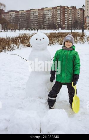 Glücklicher Junge, der in der Nähe des Schneemanns im Winterpark steht Stockfoto