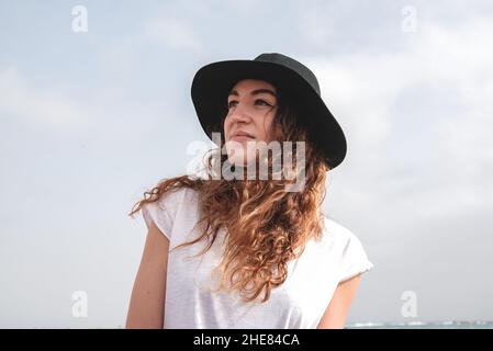 Lächelnde junge Frau in einem schwarzen Hut am Strand. Blick in die Zukunft. Stockfoto