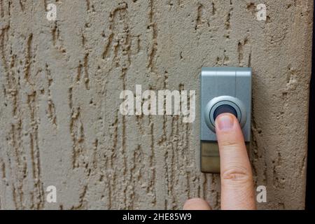 Selektiver Fokus auf einen kaukasischen Finger, der einen runden Knopf an einer Außenglocke drückt. Die Türklingel ist an einer strukturierten Wand montiert Stockfoto