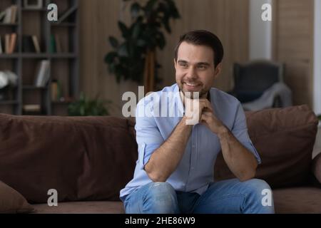 Lächelnder, verträumter Mann, der auf der Couch sitzt und in die Ferne blickt Stockfoto