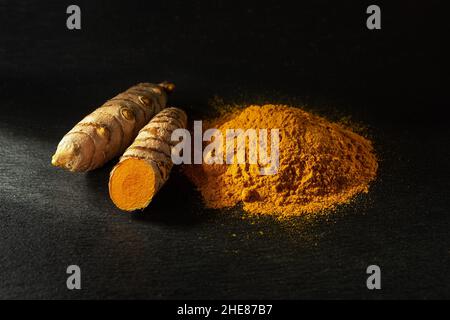 Rohe ganze Kurkuma-Wurzeln und organisches Curcuma-Pulver auf schwarzem Schiefergrund. Frische und gemahlene Rhizome von Curcuma longa linn. Indische Gewürze. Stockfoto