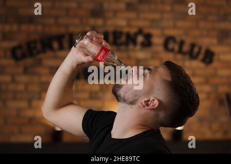 RIVNE, UKRAINE JUNI 26 2020: Junger bärtiger Mann trinkt Coca Cola aus der Flasche im Friseurladen auf Backsteinmauer Hintergrund. Stockfoto