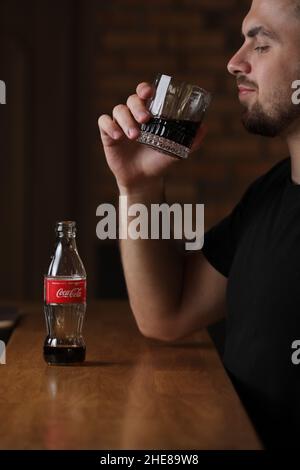 RIVNE, UKRAINE JUNI 26 2020: Junger bärtiger Mann trinkt im Café Coca Cola aus Glas. Flasche mit Cola steht auf einem Holztisch. Stockfoto