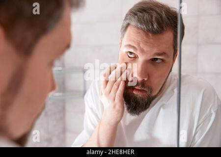 Porträt eines Mannes in weißem T-Shirt, der im Spiegel auf das Auge schaut. Gesundheitskonzept. Stockfoto