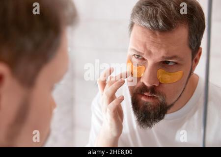 Porträt eines Mannes in weißem T-Shirt, der im Badezimmer Augenflecken auf sein Gesicht aufträgt. Self-Care-Verfahren am Morgen Stockfoto