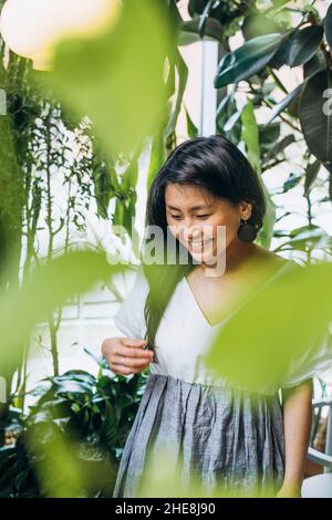 Junge Asiatin mit langen schwarzen Haaren in weiß-grauem Kleid sieht glücklich aus und lächelt unter grünen Pflanzen in veganer Café-Nahaufnahme Stockfoto