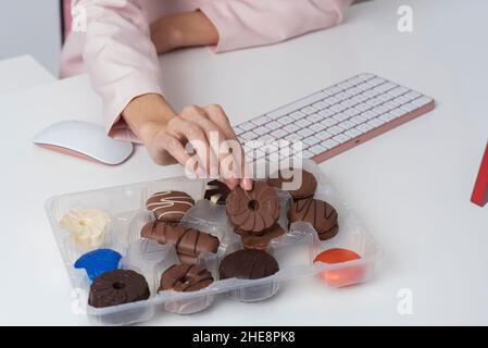 England, Großbritannien. 2021. Nahaufnahme der Hand von Frauen, die einen Schokoladenkeks aus einer Box neben einer Computertastatur nehmen. Stockfoto
