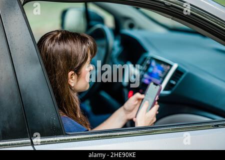 Eine kaukasische Brünette, die auf dem Beifahrersitz ihres Autos sitzt und ihr Handy anschaut Stockfoto