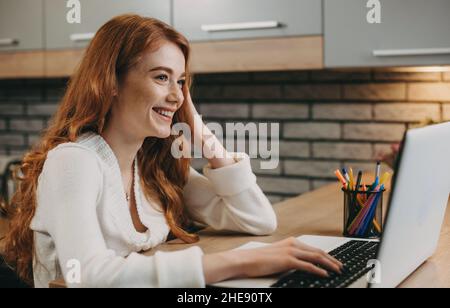 Seitenansicht einer rothaarige Frau mit Laptop-Computer, die im Heimbüro arbeitet. Internet-Technologie. Online-Kommunikation. Unternehmenstechnologie. Stockfoto