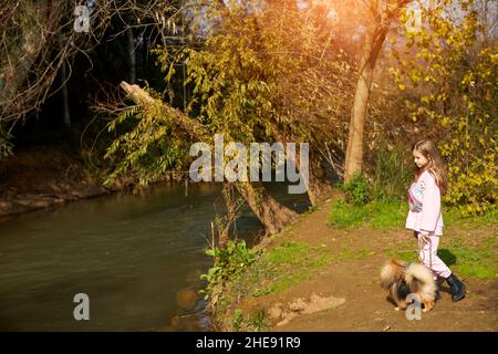 Kleines Mädchen, das den pommerschen Hund am Fluss entlang läuft
