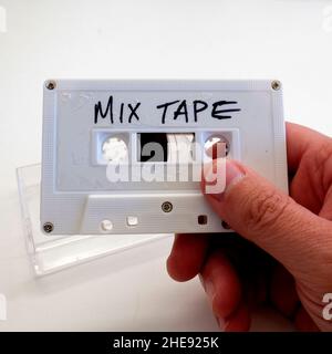 Die Hand eines Mannes hält ein weißes Kassettenband mit den Worten, DIE IN handgeschriebenen Buchstaben und einem durchsichtigen Kunststoffgehäuse im Hintergrund VERKLEBT sind. Stockfoto