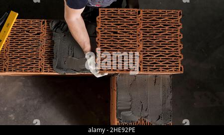 Ein Mann baut eine Backsteinmauer, Bau eines Backsteinhauses. Draufsicht auf den Industriearbeiter, der Keramikblock auf die Zementschicht legt. Stockfoto