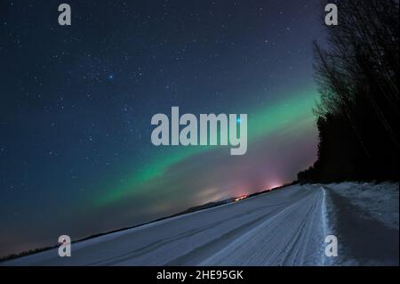 Verschneite Straße, die durch die Winterlandschaft führt. Sterne und Nordlichter am Nachthimmel. Stockfoto