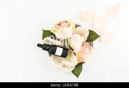 Draufsicht auf eine Schüssel mit Rosenblättern und einer Knospe mit einer Kosmetikflasche aus dunklem mattem Glas mit einem leeren Aufkleber.Unbranded mockup Stockfoto