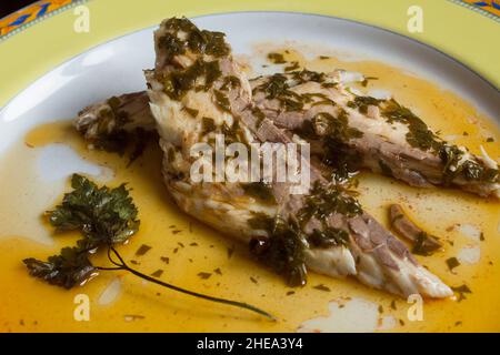 Überziehen von gebackener Seebrasse mit einem Dressing aus Knoblauchsauce, Essig, Öl und einem Zweig Petersilie in einem dekorierten, handgeschminckten Teller, der fertig zum Servieren ist. Fisch und Stockfoto