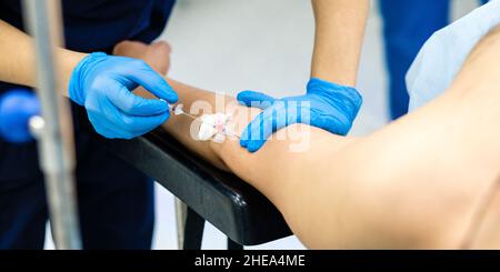 Selektiver Fokus auf die Hände eines Arztes, der eine Katheternadel in eine Vene im Arm des Patienten einführt. Die Hände des Arztes mit sterilen Handschuhen führen eine Nadel in die Hand der Person ein, um Kochsalzlösung zu injizieren. Stockfoto