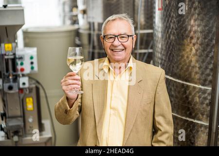 Porträt eines glücklichen älteren Mannes, dem das Weingut gehört. Er steht neben Weinlagertanks und verkostet Wein. Stockfoto