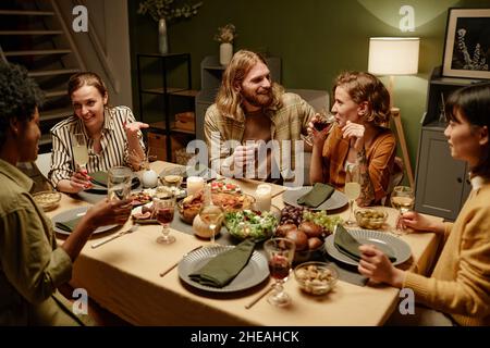 Eine Gruppe von glücklichen Freunden, die am Tisch sitzen und sich unterhalten und während der Dinner-Party im Wohnzimmer Wein trinken Stockfoto