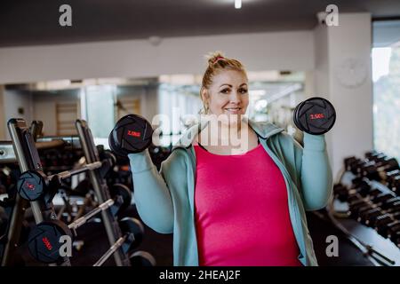 Plus size Frau Training und Heben Hanteln drinnen im Fitnessstudio, Blick auf die Kamera Stockfoto
