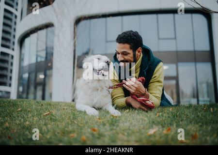 Glücklicher junger Mann, der mit seinem Hund draußen in der Stadt auf Gras liegt. Stockfoto