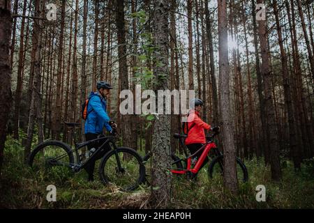 Seitenansicht von älteren Bikern, die am Herbsttag im Wald E-Bikes im Freien laufen und schieben. Stockfoto