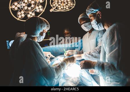 Ein internationales Ärzteteam führt eine komplexe chirurgische Operation an einem unter Anästhesie stehenden Patienten durch. Moderner Operationssaal und erfahrene Chirurgen Stockfoto