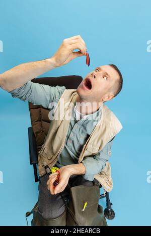 Lustiger junger Mann, Fischer mit Angelzubehör sitzt auf einem Stuhl isoliert über blauem Studiohintergrund Stockfoto