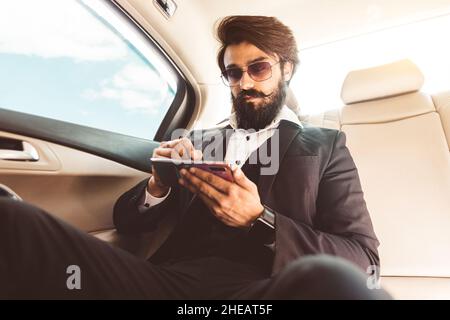 Stilvoller gutaussehender Hindu-Geschäftsmann mit Brille und Bart arbeitet auf dem Rücksitz des Autos und nutzt das Telefon. Sichere und komfortable Luxusreisen Stockfoto