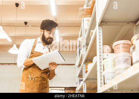 Junger attraktiver Geschäftsmann ein Töpfer mit Bart und Schnurrbart arbeitet in seiner Werkstatt. Speichert Aufzeichnungen und transkribiert sie in einem Notizbuch, das überprüft wird Stockfoto